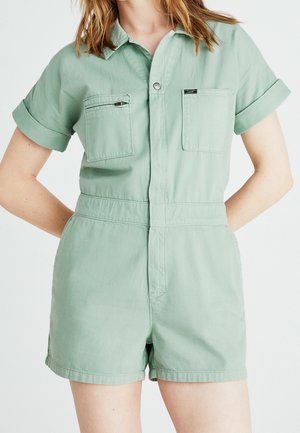 Donna con tuta corta utility verde salvia a maniche corte, con tasca con cerniera frontale e due tasche sul petto, in piedi con le mani dietro la schiena.