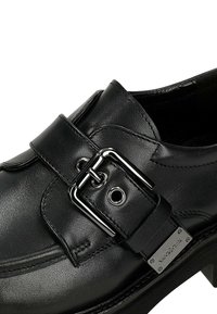Zwarte leren schoen met een rechthoekige gespsluiting en metallic accenten. Glad textuur met een iets verhoogde, solide hak.