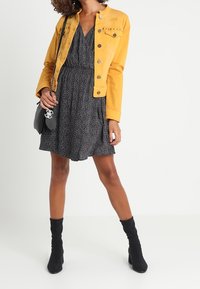 Žena v žluté zapínací bundě přes černé vzorované šaty, držící černou kabelku přes rameno a obutá v černých botách do poloviny lýtek.