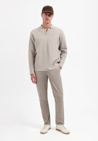 Beige polo shirt met lange mouwen en een relaxte pasvorm, met een V-hals kraag, gecombineerd met lichtbeige slim-fit broek en witte sneakers. Bruine pet.