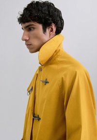 Chaqueta amarilla con cuello alto, que presenta cierres de toggles negros y una textura suave. Las mangas son de corte relajado, destacando la simplicidad.