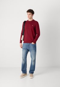 Bordeaux genser med rund hals og ribbestrikkede mansjetter, sammen med lyseblå rette bukser og beige joggesko. Modellen holder en liten beige veske.