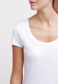 Bílý bavlněný tričko s kulatým výstřihem a krátkými rukávy, s hladkou texturou a přiléhavým střihem. Minimalistický styl.