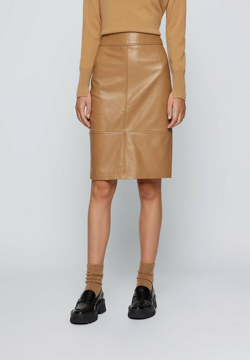 Femme portant une jupe en cuir couleur camel jusqu'aux genoux, un pull assorti camel, des chaussettes camel et des mocassins noirs épais, debout devant un fond uni.