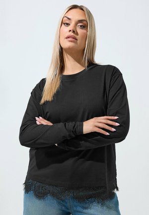 Blondynka z prostymi włosami, ubrana w czarną bluzkę z długim rękawem z koronkowym wykończeniem oraz niebieskie dżinsy, stojąca z założonymi ramionami.