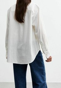 Vit skjorta med avslappnad passform, som har långa ärmar, rundad nederkant och en strukturerad tygkvalitet. Kombineras med mörkblå vida jeans.