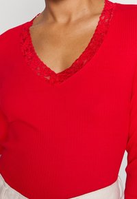 Punainen ribattu pusero V-päällä, jossa on pitsireuna. Kangas on sileä ja tiukka, korostaen kaula-aukkoa ja olkapäitä.