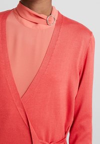 Cardigan en corail avec un décolleté en V profond, texture tricot légère, et une couche intérieure montée pêche assortie avec un accent en anneau.