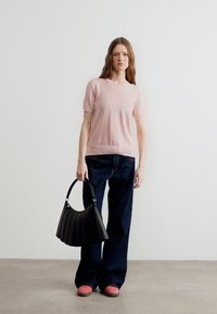 Γυναίκα με μακριά καστανά μαλλιά που φοράει ροζ ανοιχτό κοντομάνικο πουλόβερ, σκούρο φαρδύ παντελόνι τζιν, ροζ παπούτσια, κρατώντας μια μαύρη τσάντα στο πλευρό της.