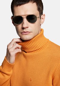 Pomarańczowy sweter z kołnierzem typu golf, prą�żkowany, z małym haftem. Modelka nosi okulary przeciwsłoneczne w metalowej oprawie z ciemnymi szkłami.