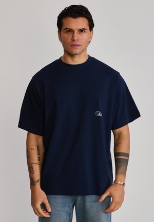 Uomo con capelli corti e scuri che indossa una maglietta oversize blu navy con piccolo logo bianco e jeans blu, in piedi contro uno sfondo uniforme.