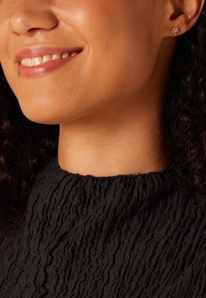 Boucle d'oreille en forme d'arbre en or portée par une femme aux cheveux bouclés, vêtue d'un haut en tissu noir texturé avec un col montant. La texture douce de sa peau est visible.