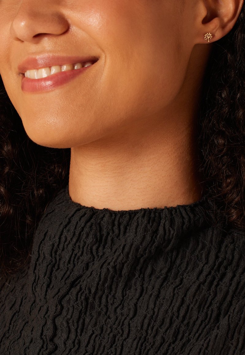 Boucle d'oreille en forme d'arbre en or portée par une femme aux cheveux bouclés, vêtue d'un haut en tissu noir texturé avec un col montant. La texture douce de sa peau est visible.
