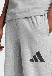 Sivé tepláky vyrobené z mäkkej látky, s elastickým pásom, bočnými vreckami a čiernym logom Adidas na ľavom stehne.