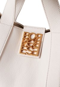 Weiße Ledertragetasche mit einem auffälligen goldenen Logo-Plakette mit einem Ausschnittdesign an der Vorderseite. Strukturierte Oberfläche mit Steppdetails.