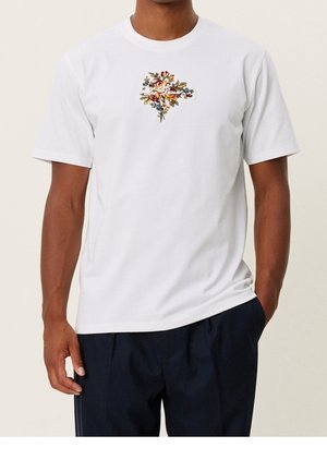 Homme portant un t-shirt blanc avec un petit bouquet floral coloré centré sur la poitrine, associé à un pantalon foncé.