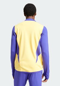 Chaqueta deportiva de manga larga en amarillo y púrpura, con un patrón texturizado en los hombros y los costados, diseñada para un ajuste ceñido.