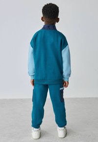 Sudadera color teal con mangas y cuello azul claro, pantalones de chándal teal a juego, que presentan bolsillos laterales y zapatillas blancas con el texto "BAKER".