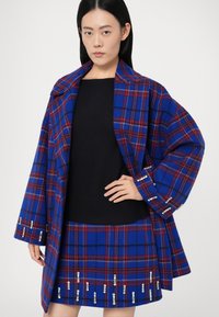 Casaco em padrão xadrez azul e vermelho com um colarinho largo, corte descontraído, e detalhes em joia nas mangas. Combinado com uma blusa preta texturada e uma saia xadrez.