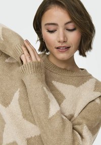 Licht beige gebreide trui met sterpatronen in een lichtere tint, voorzien van geribde manchetten en een ronde halslijn. Zachte textuur, gemiddeld gewicht.