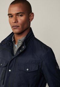 Chaqueta impermeable de color azul marino con un cuello alto, detalles de botones a presión, dos bolsillos en el pecho y un acabado de tejido texturizado.