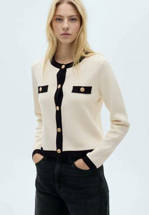 Cream cardigan with black trim, featuring gold buttons and two front pockets. The fabric appears soft with a ribbed texture at the edges.