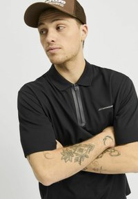 Zwarte polo met korte mouwen, ritskraag en een subtiele applicatie op de borst. Tan pet met logo op een bruine mesh achterkant.