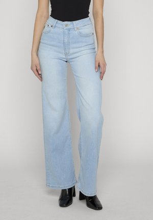 Kvinne modell iført lyseblå høytlivs vide bukser, svart topp og svarte ankelstøvler med hæl, stående mot en ensfarget lys bakgrunn.