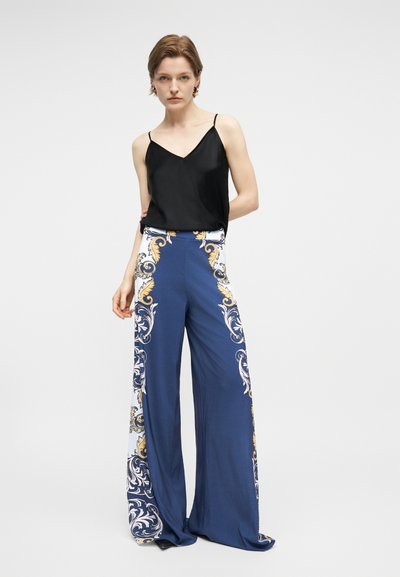 Γυναίκα που στέκεται με τα χέρια πίσω από την πλάτη, φορώντας μαύρο αμάνικο τοπ και φαρδιά μπλε παντελόνια με λευκό και χρυσό μπαρόκ μοτίβο.