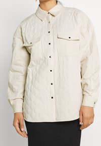Chaqueta de camisa acolchada en color beige claro con cuello, dos bolsillos en el pecho y botones de presión plateados. Presenta un patrón de diamante y una textura suave.