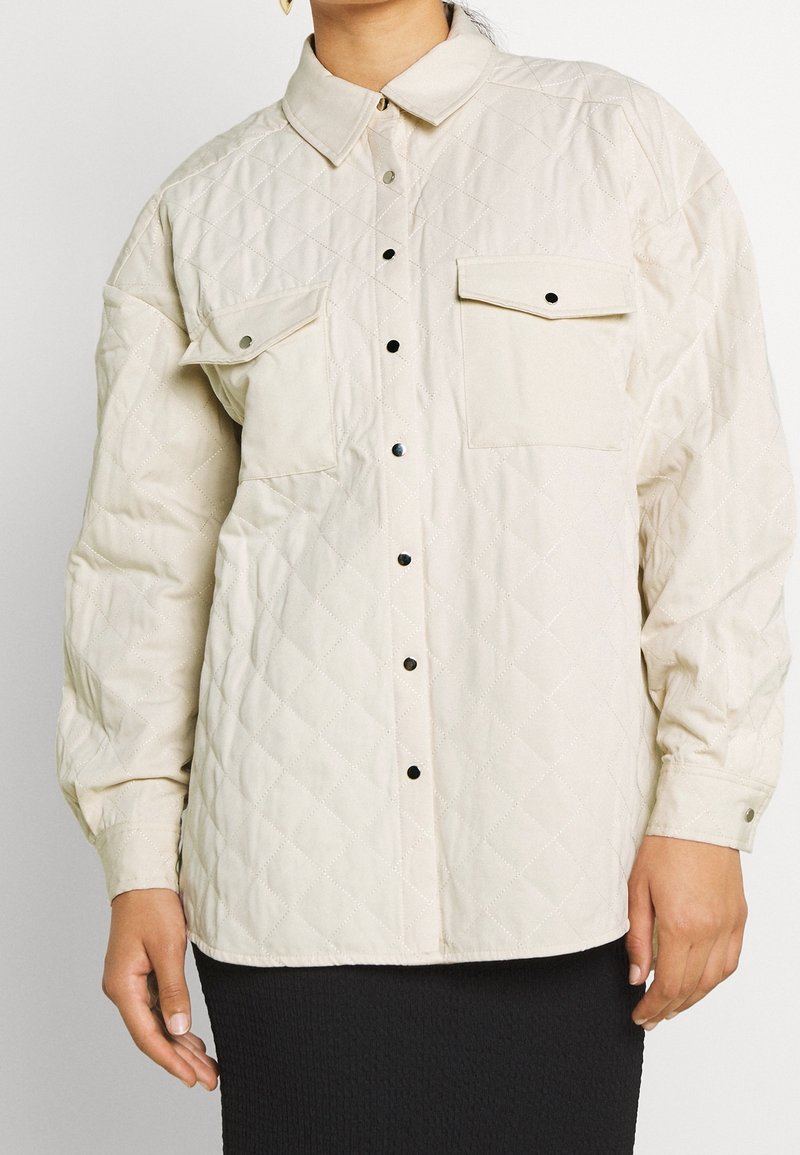 Chaqueta de camisa acolchada en color beige claro con cuello, dos bolsillos en el pecho y botones de presión plateados. Presenta un patrón de diamante y una textura suave.