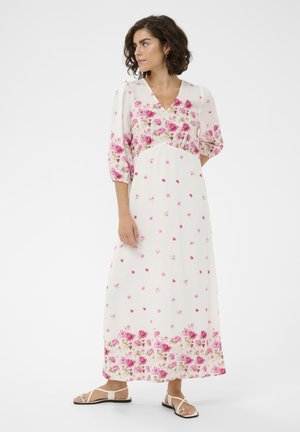 Femme aux cheveux bouclés portant une robe longue blanche à motifs floraux roses et des sandales blanches, debout et regardant vers sa gauche.