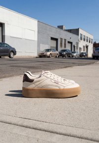 Λευκό πλατφόρμα sneaker με καφέ σόλα τοποθετημένος σε πεζοδρόμιο της πόλης, με αυτοκίνητα και βιομηχανικά κτίρια στο φόντο κάτω από καθαρό ουρανό.