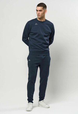 Vīrietis tumši zilā sporta džemperī un atbilstošās biksēs stāv ar rokām aiz muguras, valkājot baltas sporta kurpes, vienkārša gaiša fona priekšā.