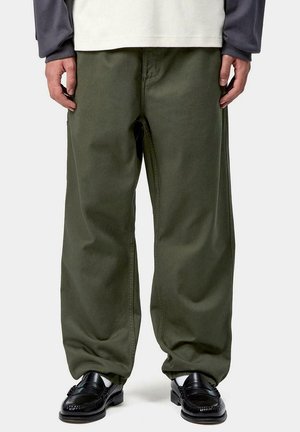 Persona che indossa pantaloni larghi verde oliva, mocassini in pelle nera, calzini bianchi e una maglietta a maniche lunghe bianca e grigia, in piedi su uno sfondo bianco.