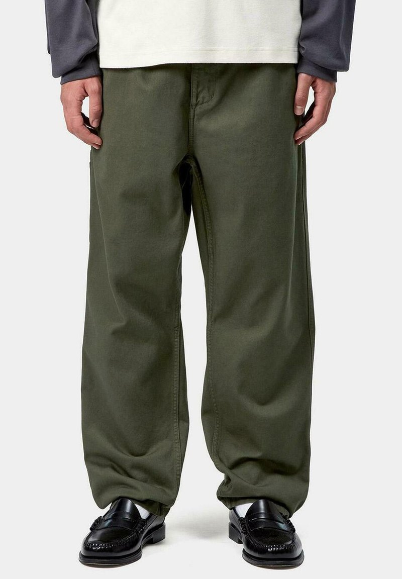 Persona con pantalones holgados de color verde oliva, mocasines de cuero negros, calcetines blancos y una camisa de manga larga blanca y gris, de pie sobre un fondo blanco.