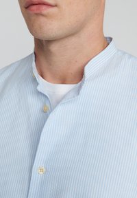 Lichtblauwe gestreepte shirt met een bandkraag, voorzien van een voorflap met beige knopen. De stof lijkt glad en heeft een lichte textuur.