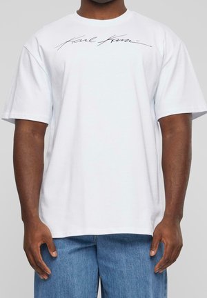 Weißes Baumwoll-T-Shirt mit kurzen Ärmeln, das ein schwarzes Signaturdesign über der Brust aufweist. Klassische Rundhalslinie und lockere Passform.