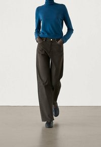 Person wearing blue turtleneck sweater, dark wide-leg pants, and textured dark slip-on shoes, walking with hands in pockets on neutral floor.