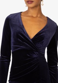 Navyblaues Samtkleid mit tiefem V-Ausschnitt, langen Ärmeln und gerafftem Detail an der Taille, das eine glatte und weiche Textur zeigt.