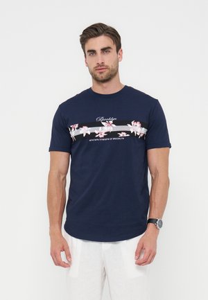 Bărbat purtând un tricou albastru închis cu model floral și textul „Brooklyn”, asortat cu pantaloni albi și un ceas de mână, stând pe un fundal alb.