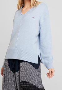 Pull en maille bleu clair avec un col en V et un ourlet côtelé. Il présente des manches longues et des fentes latérales pour plus de liberté de mouvement. Détail logo discret visible.