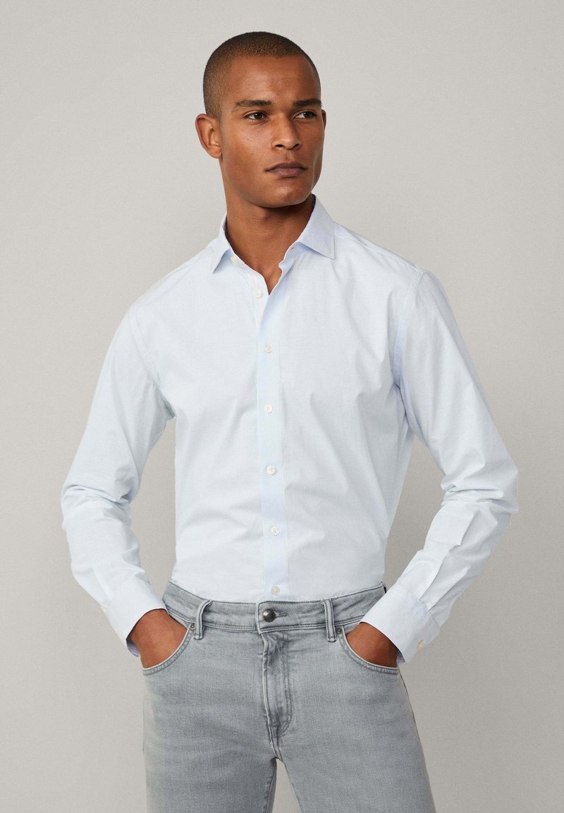 Light blue long-sleeve shirt with button-down collar, cotton fabric, structured fit, worn with grey jeans, displaying a simple, clean design.