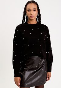 Pull en maille noir avec des détails en perles, col rond et manches longues, associé à une jupe noire en simili cuir ornée de perles.