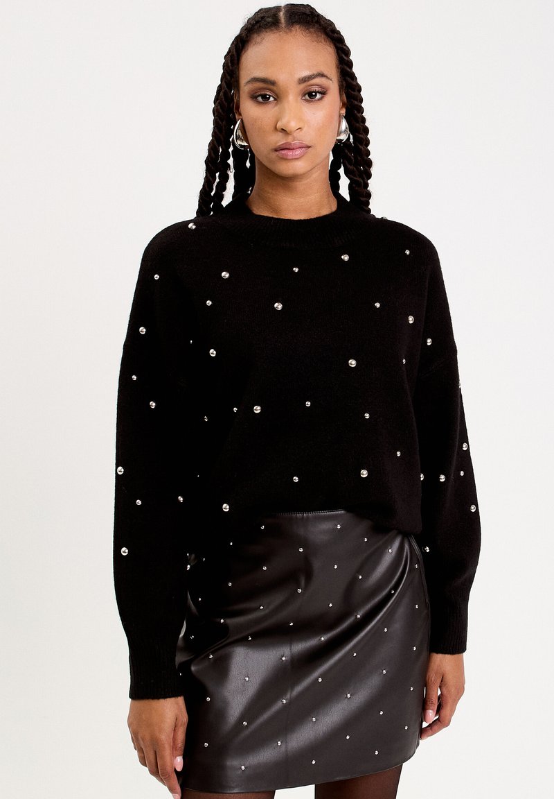 Pull en maille noir avec des détails en perles, col rond et manches longues, associé à une jupe noire en simili cuir ornée de perles.