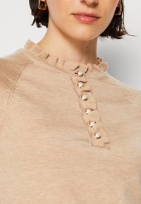 Femme portant un haut en maille beige avec un col volanté et six boutons en perles sur le devant, avec des boucles d'oreilles créoles dorées.