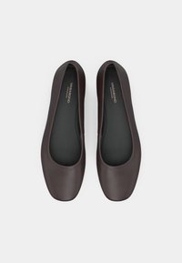 Un paio di ballerine in pelle marrone scuro con punta quadrata arrotondata e soletti neri etichettati "Vagabond Shoemakers" su uno sfondo bianco.