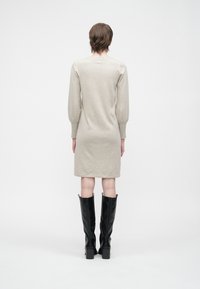 Oseba s kratkimi rjavimi lasmi, oblečena v bež dolg rokav do kolen, z črnimi visokimi čevlji do kolen, stoji obrnjena stran od bele ozadje.