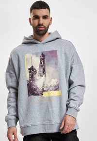 Sudadera gris con capucha que presenta un gráfico de un lanzamiento de cohete y el texto "EN MOVIMIENTO". Mezcla de algodón, corte relajado, textura suave.