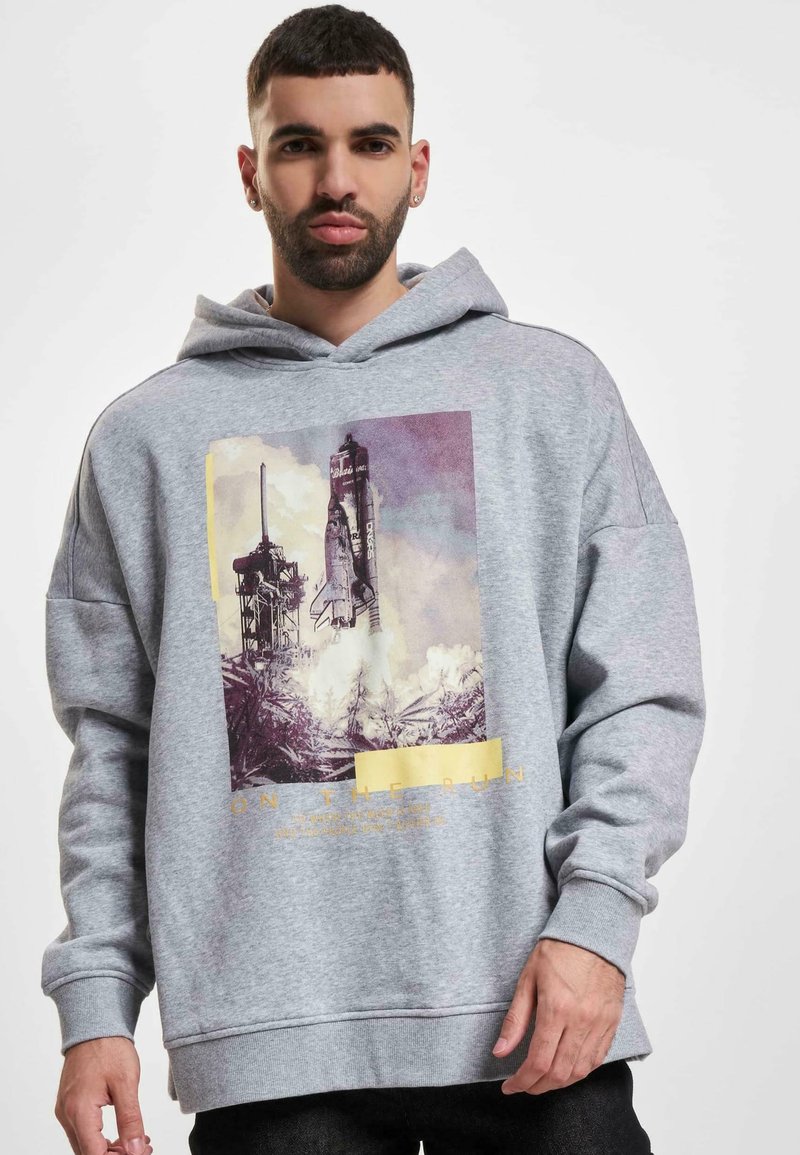 Sudadera gris con capucha que presenta un gráfico de un lanzamiento de cohete y el texto "EN MOVIMIENTO". Mezcla de algodón, corte relajado, textura suave.